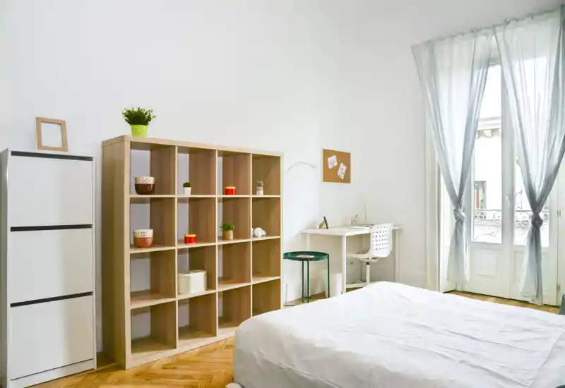Chambre lumineuse et minimaliste avec un lit confortable, une étagère en bois, un petit bureau et une grande fenêtre apportant une belle lumière naturelle — aérée et fonctionnelle pour les locations.