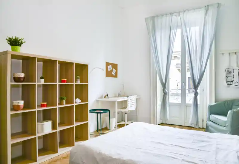Chambre lumineuse et aérée avec lit bien fait, un bureau près de la fenêtre et une étagère modulable pour le rangement — parfaite pour un séjour confortable avec lumière naturelle.