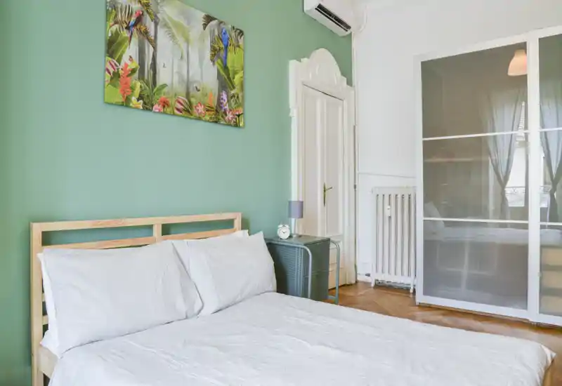 Chambre lumineuse et bien mise en scène avec un cadre de lit en bois clair, linge de lit blanc et un mur vert orné d'une toile colorée. On note une table de chevet avec lampe et une armoire aux panneaux dépoli.