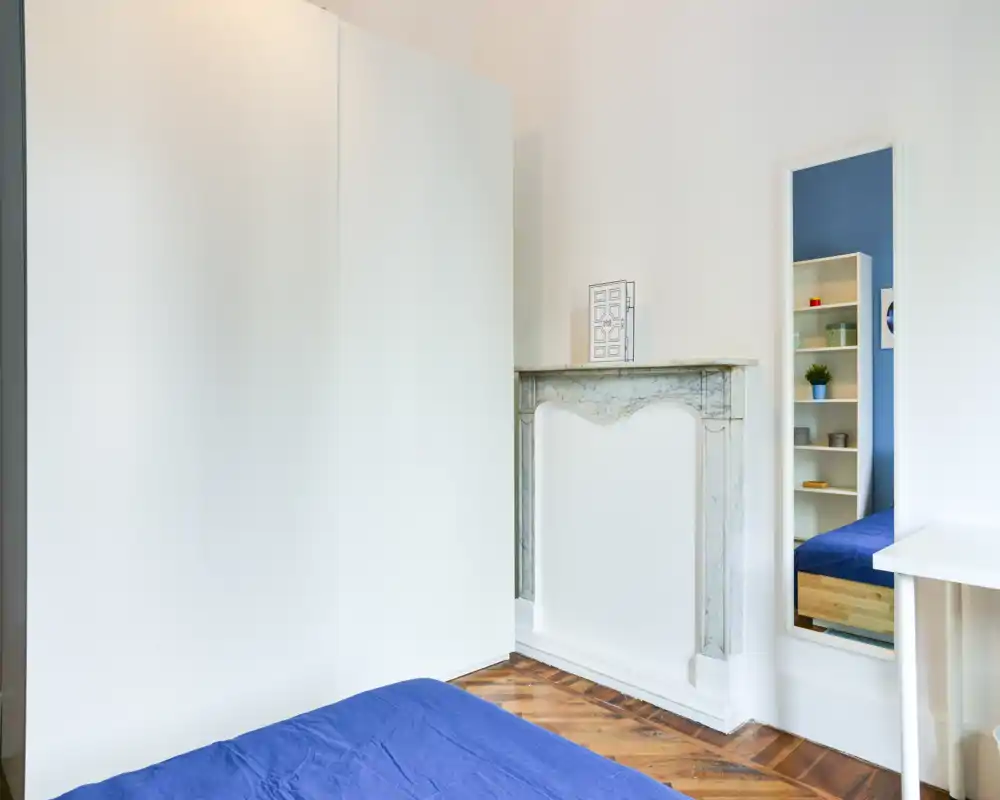 Camera da letto luminosa e minimale con letto con copriletto blu, armadio bianco e un caminetto decorativo; lo specchio riflette una libreria aumentando la profondità.