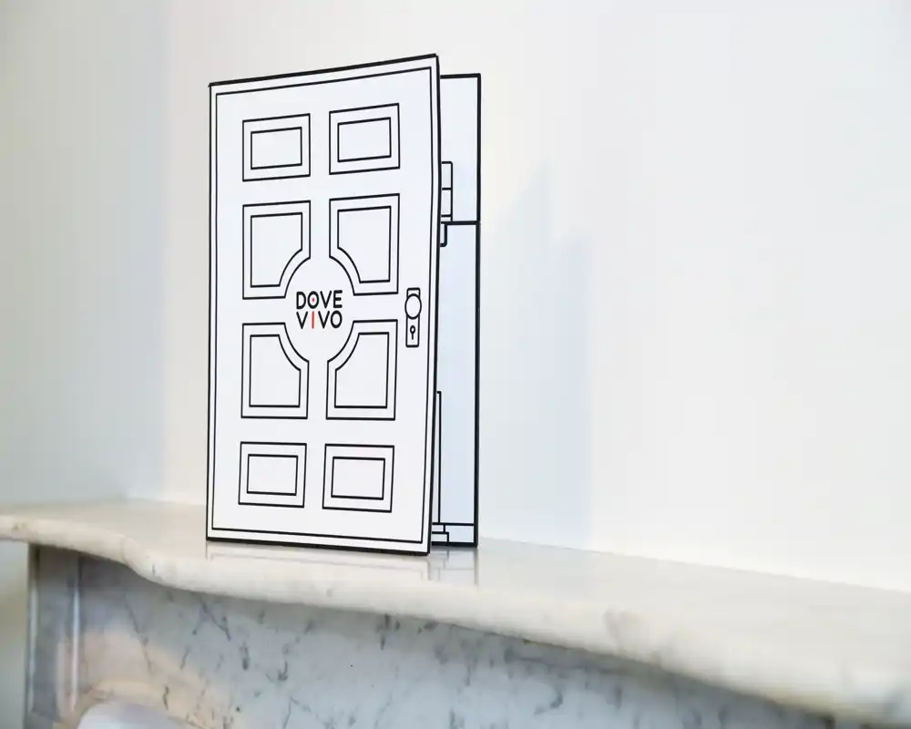 Primo piano di un cartoncino/oggetto decorativo a forma di porta appoggiato su un caminetto in marmo davanti a una parete bianca. L'immagine mette in risalto l'oggetto e la superficie, non la planimetria o la funzione di una stanza.