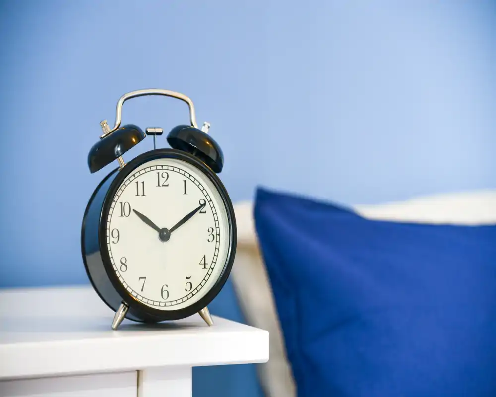 Primo piano di un comodino con una sveglia nera classica e parte di un cuscino blu e testiera visibili, che suggeriscono un angolo camera da letto luminoso e dallo stile semplice.