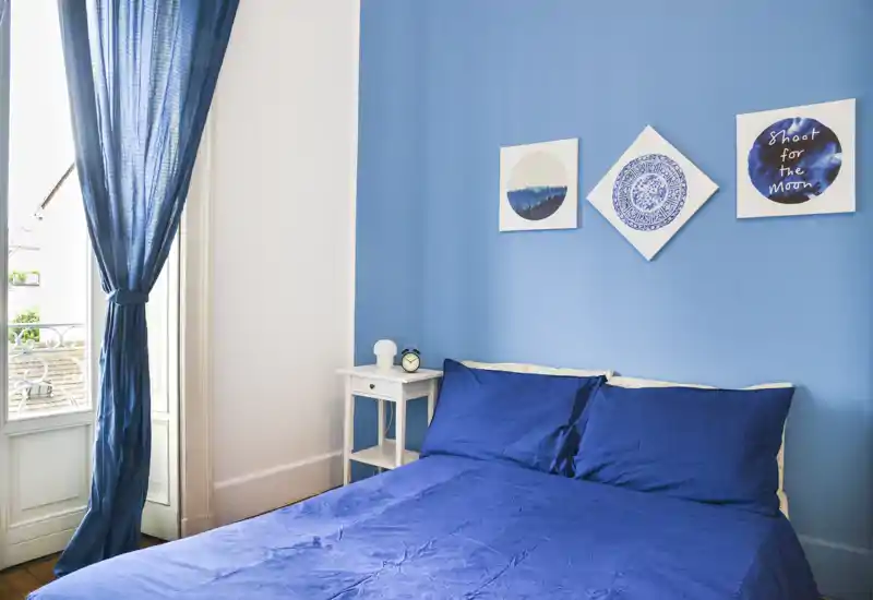 Chambre lumineuse aux tons bleus avec lit double, linge coordonné, tableaux au-dessus du lit, table de chevet et lumière naturelle depuis une porte-fenêtre.