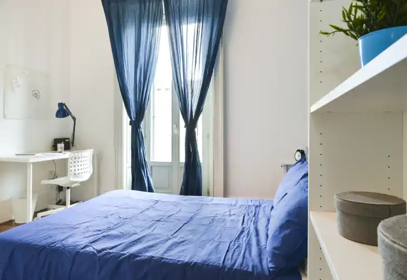 Chambre lumineuse et soignée avec lit double en linge bleu, bureau avec lampe près de la fenêtre et étagère avec boîtes de rangement décoratives.