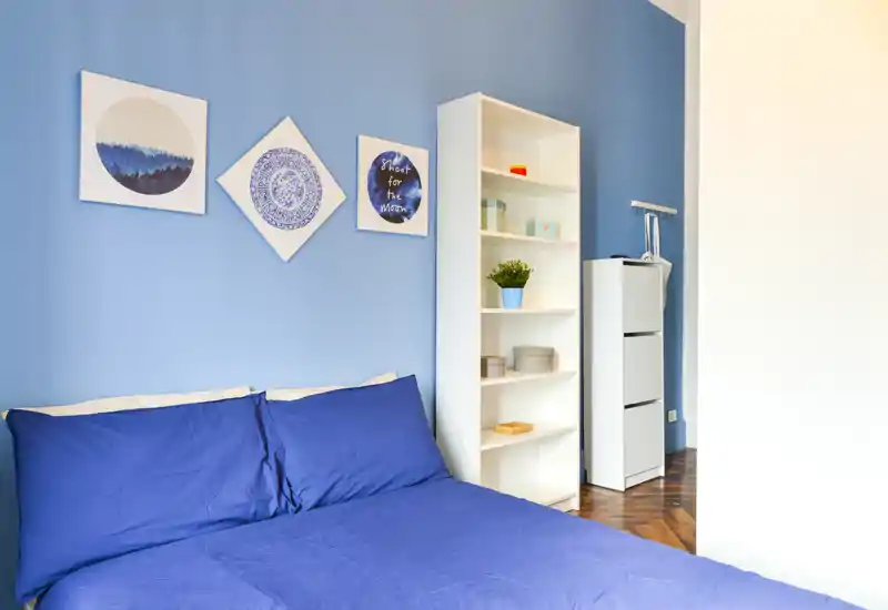 Chambre lumineuse et bien rangée avec un mur bleu, un lit double en linge bleu foncé; étagères simples et tableaux apportent une ambiance moderne et chaleureuse.