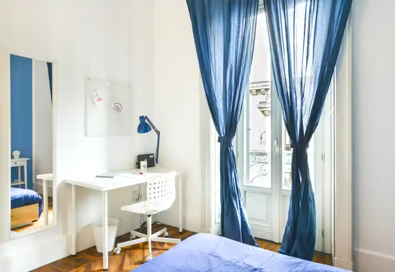 Chambre lumineuse et moderne aux accents bleus : grande fenêtre avec rideaux voile bleu marine, lit bien fait et bureau blanc avec lampe de travail — idéale pour un étudiant ou une personne seule.