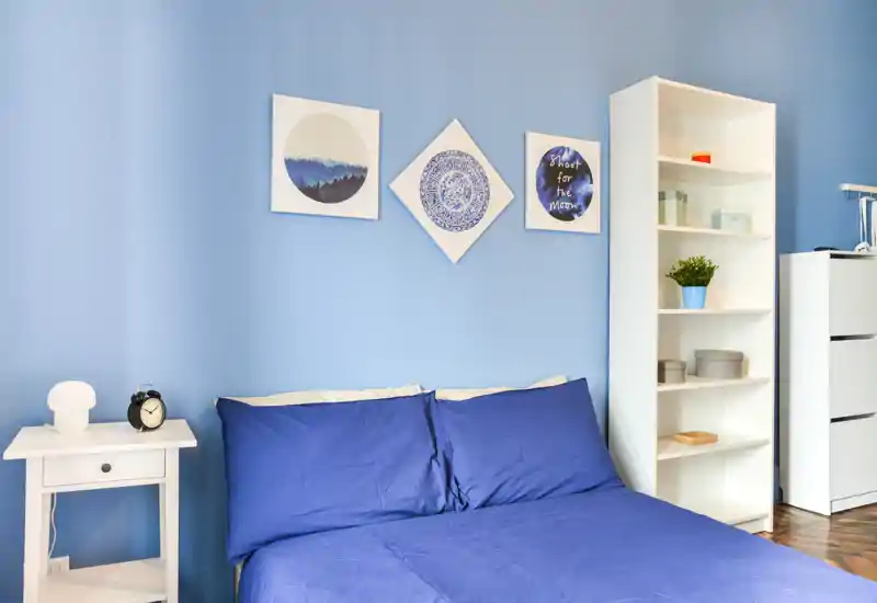 Chambre lumineuse et soignée avec un lit double en linge bleu foncé, des tableaux assortis et une table de chevet et une étagère blanches pour le rangement.
