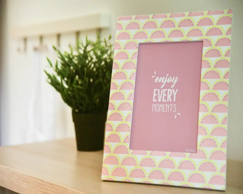 Gros plan d'un cadre photo décoratif avec un insert rose indiquant « enjoy EVERY MOMENTS », posé sur une surface en bois à côté d'une petite plante en pot. L'image met l'accent sur un détail de décoration plutôt que sur une pièce identifiable.