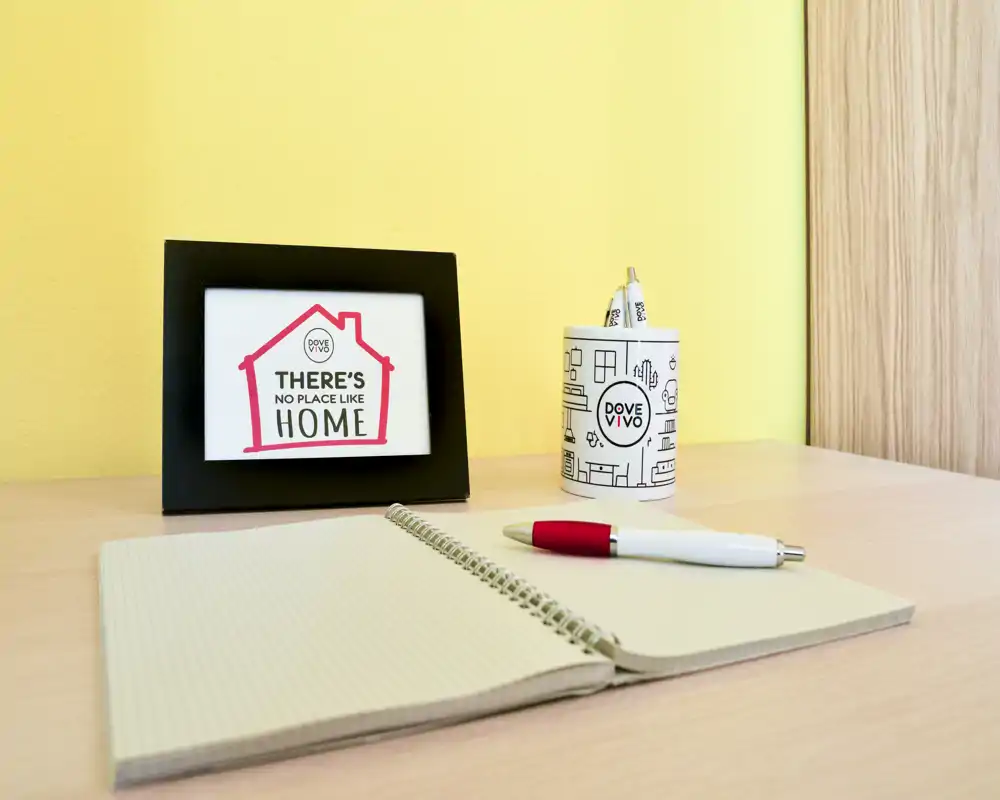 Petit coin bureau soigné avec un carnet ouvert, un stylo et un cadre décoratif sur un mur jaune pâle — un espace de travail compact adapté aux notes et petites tâches.