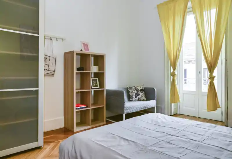 Chambre lumineuse et bien rangée avec un grand lit au premier plan, un petit canapé confortable, une étagère à cubes et de hautes fenêtres avec rideaux jaunes laissant entrer beaucoup de lumière naturelle.