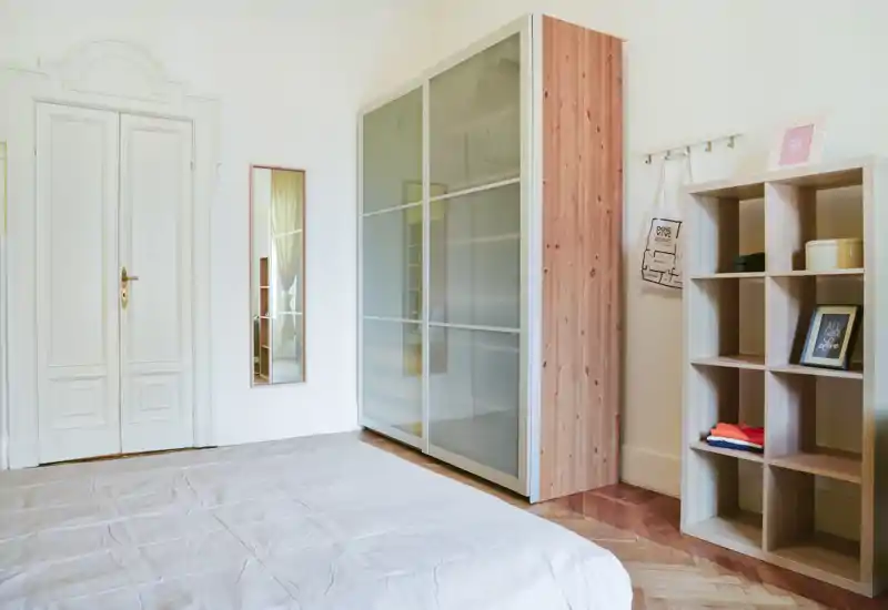 Chambre lumineuse et minimaliste avec un lit bas bien fait, une grande armoire aux portes dépoli et une étagère à compartiments. La lumière naturelle et le parquet en bois apportent une ambiance chaleureuse et aérée.
