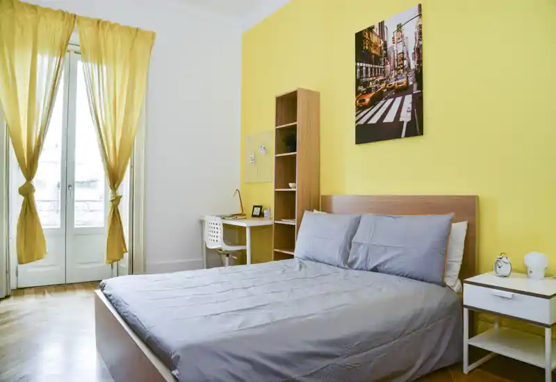 Chambre lumineuse et moderne avec un lit double contre un mur jaune, table de chevet et un petit bureau près de grandes fenêtres à la française offrant une belle lumière naturelle.