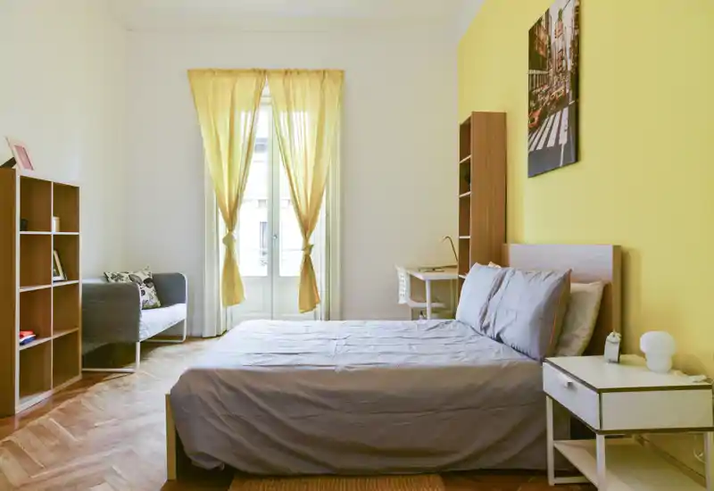 Chambre lumineuse et soignée avec lit double, table de chevet et bureau près d'une grande fenêtre aux rideaux jaunes. Parquet en bois, petit canapé et étagères pour le rangement — un espace accueillant et bien éclairé.