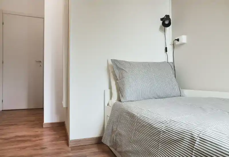 Chambre lumineuse et minimaliste avec un lit simple habillé de linge rayé et une lampe de lecture murale — lignes épurées et sol en bois pour une ambiance moderne et apaisante.