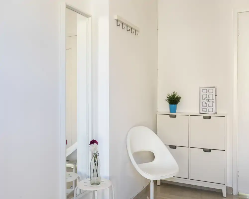 Entrada minimalista y luminosa con una silla blanca moderna, perchas en la pared, un espejo estrecho y un pequeño armario para zapatos — zona de bienvenida ordenada.