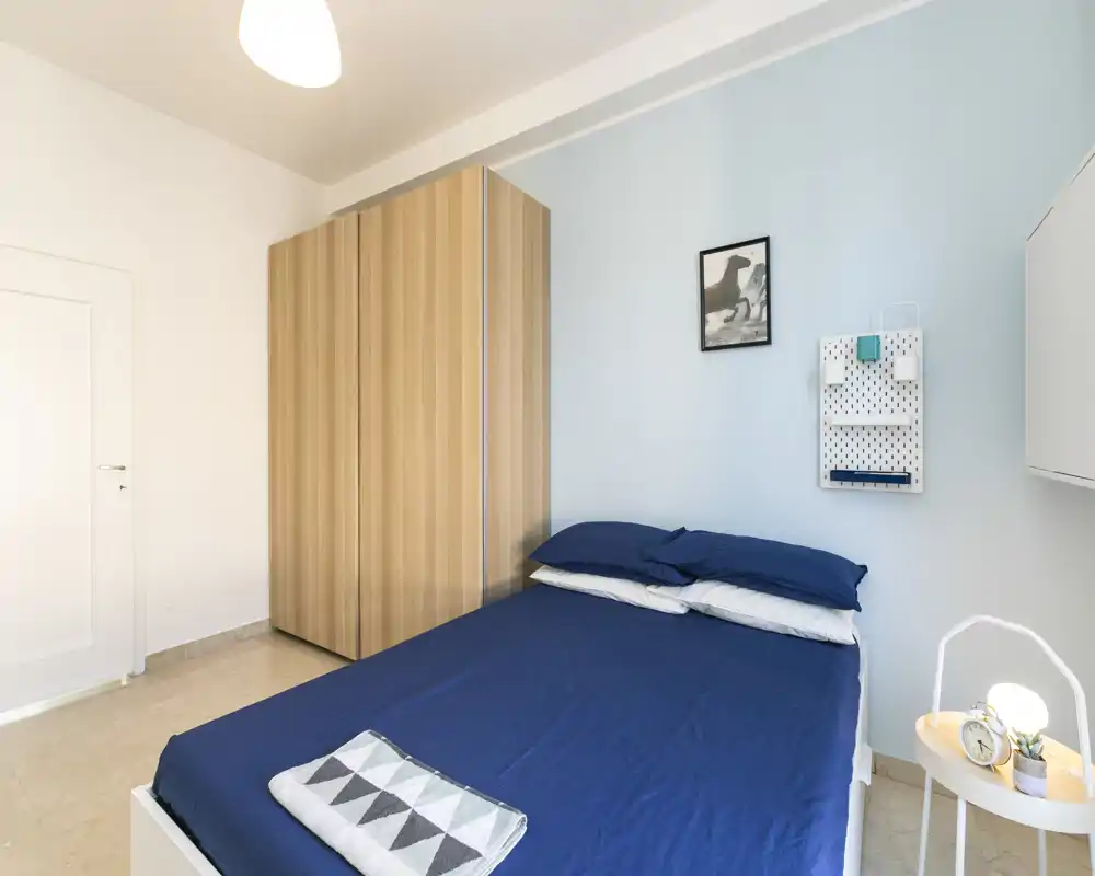 Dormitorio moderno y luminoso con cama doble con ropa de cama azul oscuro, armario de madera y mesita con lámpara — estilo limpio y minimalista perfecto para anuncios.