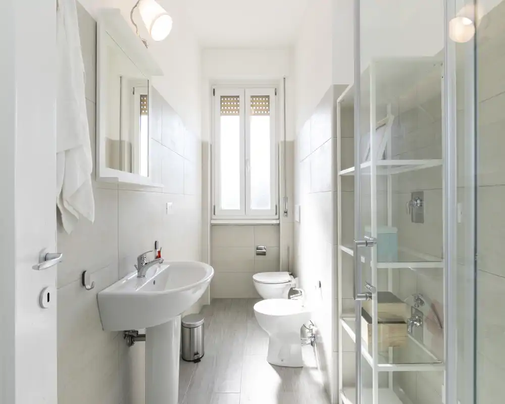 Baño moderno y luminoso con lavabo de pie, inodoro, bidé y cabina de ducha de cristal; azulejos neutros y luz natural desde la ventana generan una sensación limpia y fresca.