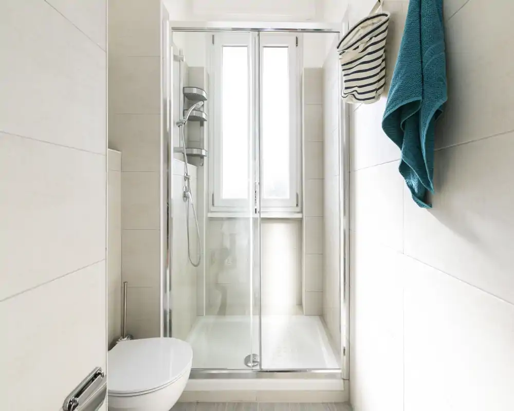 Baño moderno y luminoso con ducha acristalada, inodoro suspendido y azulejos claros en las paredes — diseño limpio y funcional.