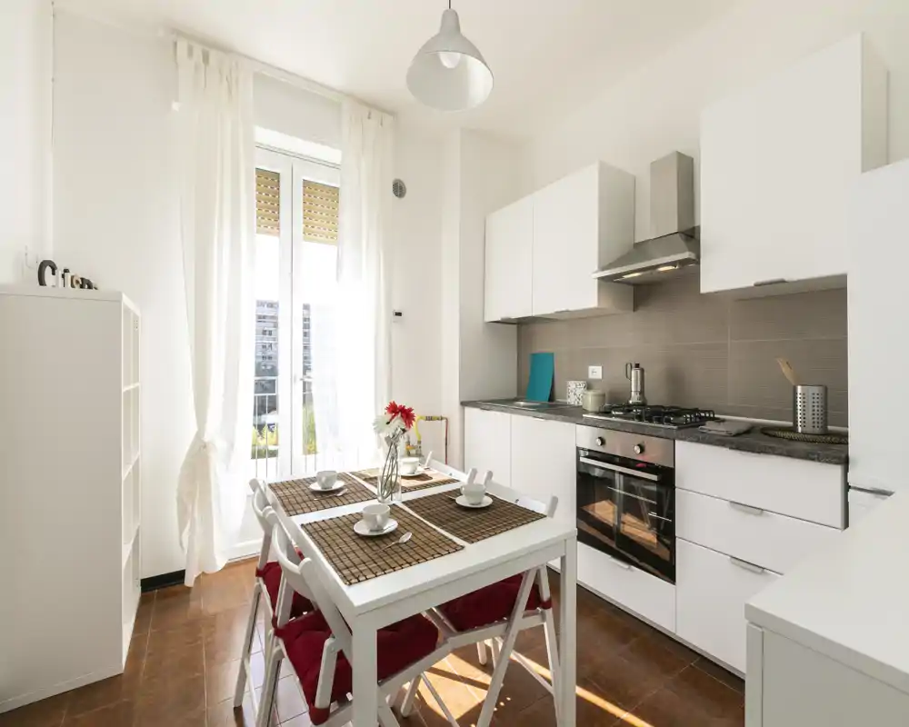 Cocina moderna y luminosa con armarios blancos, cocina de gas y una mesa comedor para cuatro junto a una ventana alta que inunda la estancia de luz natural.