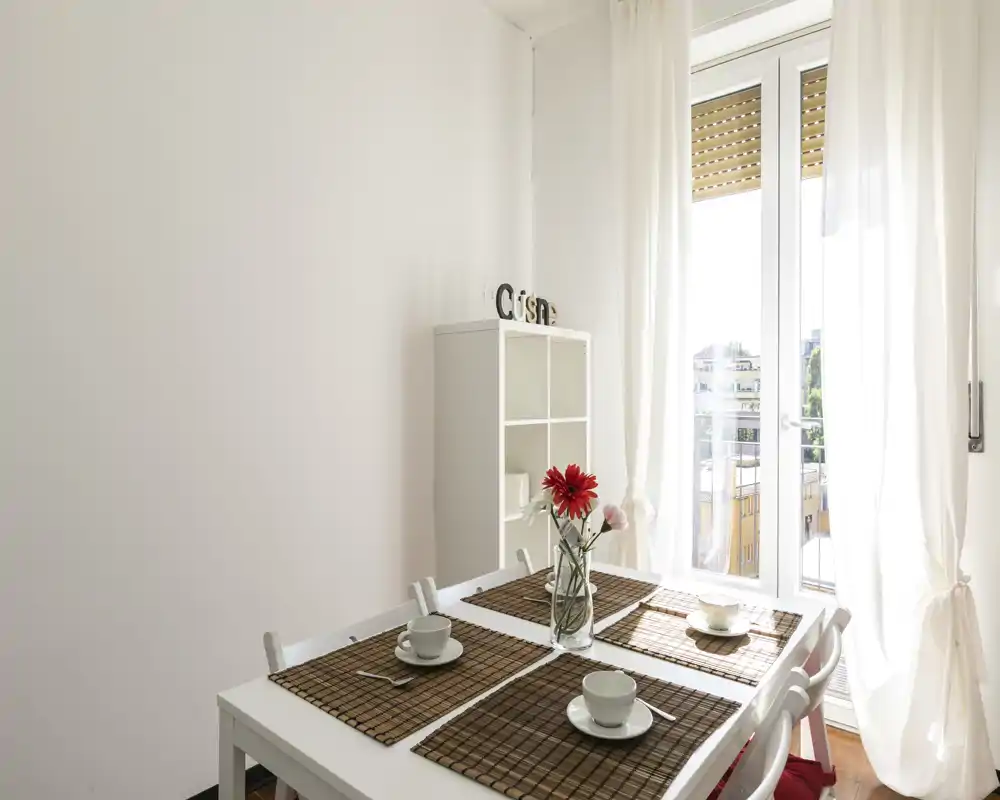 Área de comedor luminosa y minimalista con mesa blanca para cuatro, manteles individuales de mimbre y un jarrón con flores junto a una amplia puerta al balcón iluminada.