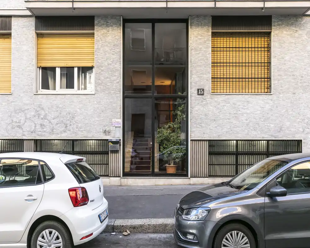 Vista a pie de calle de la entrada de un edificio residencial con puertas de cristal, escalera visible y macetas; dos coches aparcados delante.