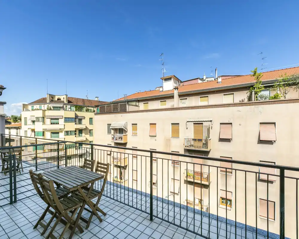 Balcón privado soleado con mesa y sillas exteriores, barandilla y vistas a edificios cercanos y cielo azul — ideal para el café matutino.