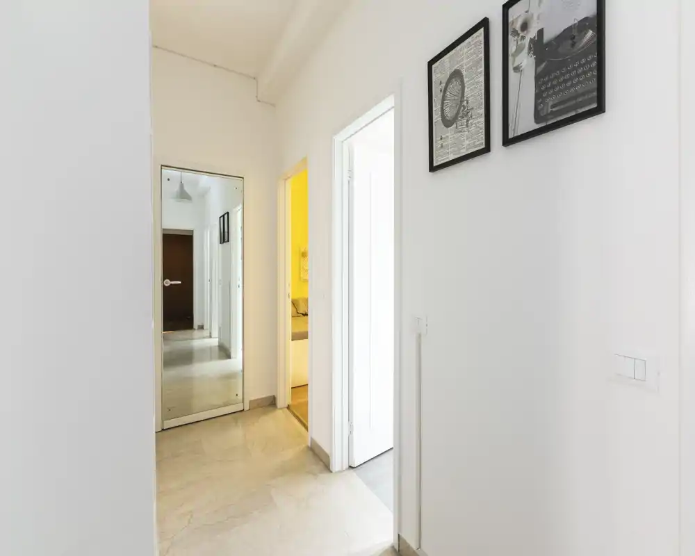 Entrada/pasillo brillante y minimalista con paredes blancas, suelo de baldosas y cuadros en la pared; un espejo y puertas abiertas conectan con las habitaciones contiguas.