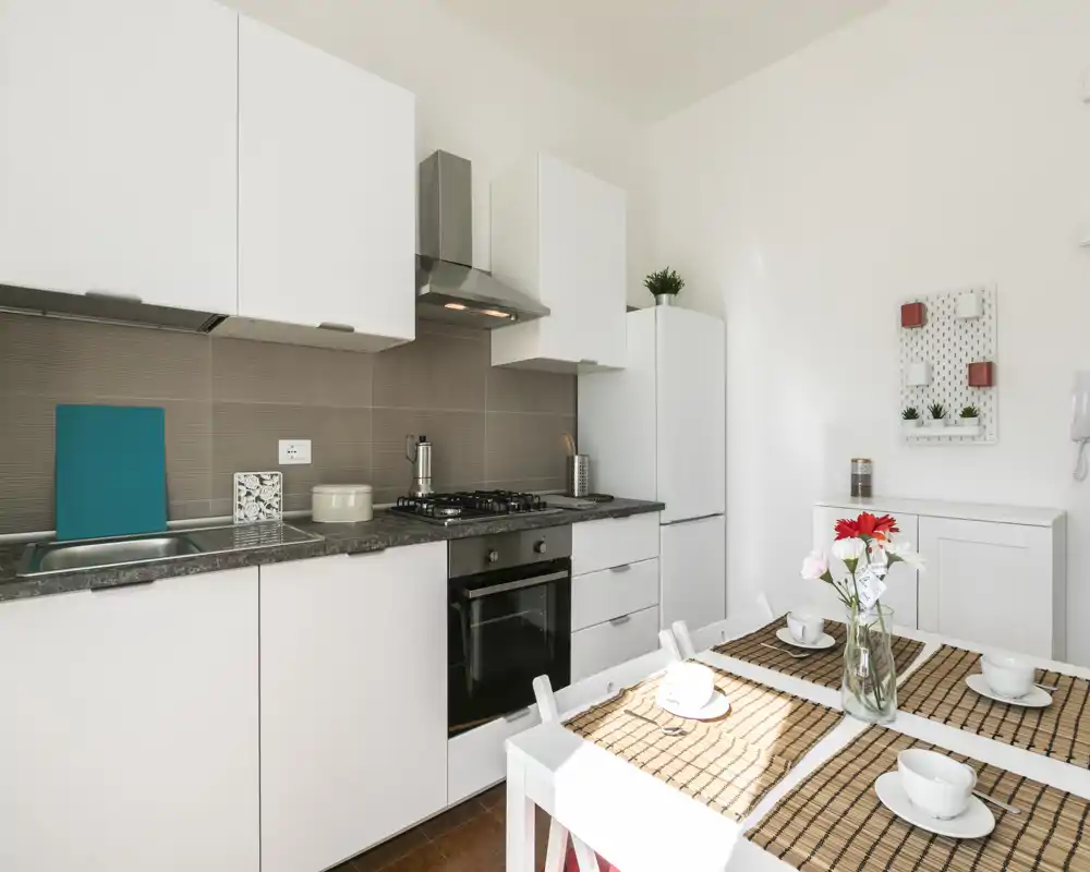 Cocina moderna y luminosa con armarios blancos, vitrocerámica de gas y mesa para cuatro; toques decorativos sencillos (flores y individuales) crean un ambiente acogedor.