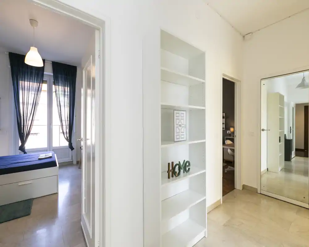Pasillo luminoso que muestra un dormitorio a través de la puerta abierta: cama individual arreglada con cortinas oscuras y luz natural procedente de la ventana.
