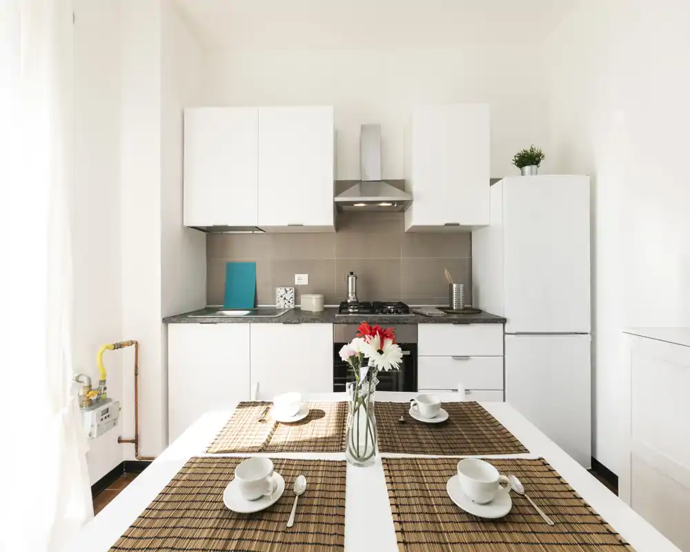 Cocina moderna y luminosa con muebles blancos, cocina de gas, frigorífico y mesa dispuesta con manteles individuales y flores frescas.