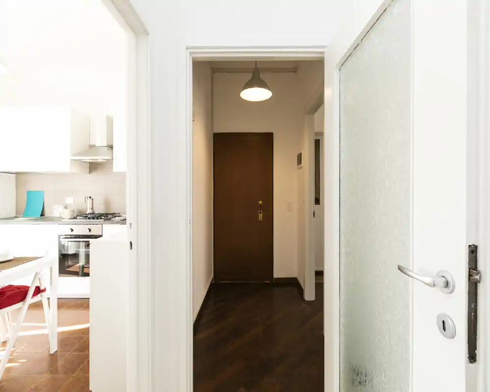 Entrada luminosa y limpia con puerta principal de madera en frente y vista parcial de la cocina contigua; puertas blancas modernas y suelo de parquet que transmiten buena acogida.