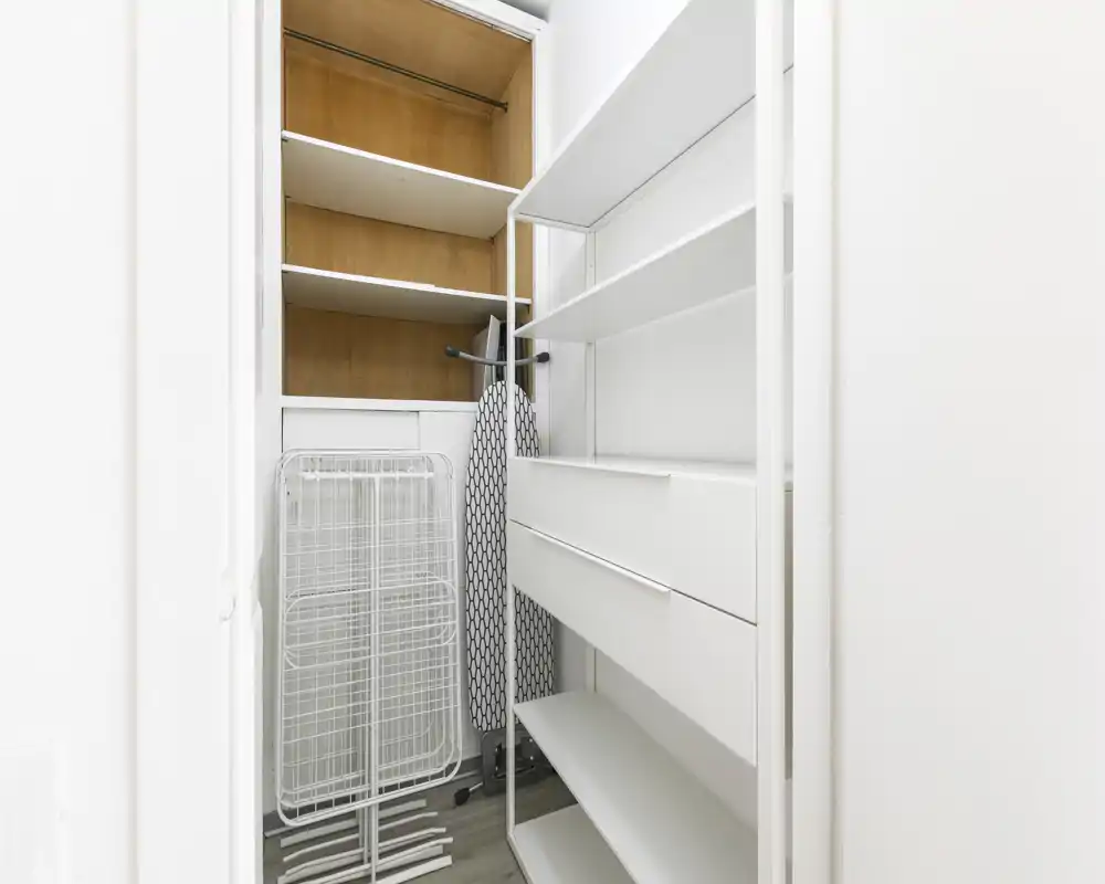 Pequeño armario/almacén de entrada con estanterías, cajones y espacio para colgar; acabado blanco y tabla de planchar guardada en su interior.