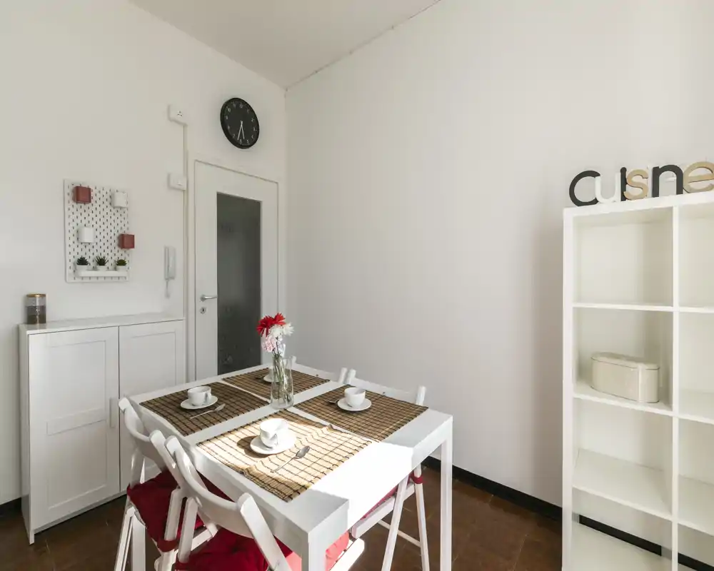 Área de comedor luminosa y minimalista con mesa blanca para cuatro, manteles decorativos y un jarrón con flores: limpia, moderna y acogedora.