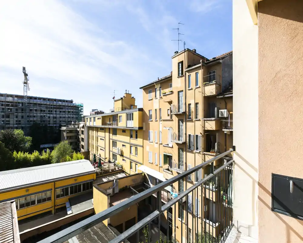 Balcón soleado de apartamento con vistas a edificios vecinos y un pequeño patio — espacio exterior luminoso con barandilla metálica y agradable vista urbana.