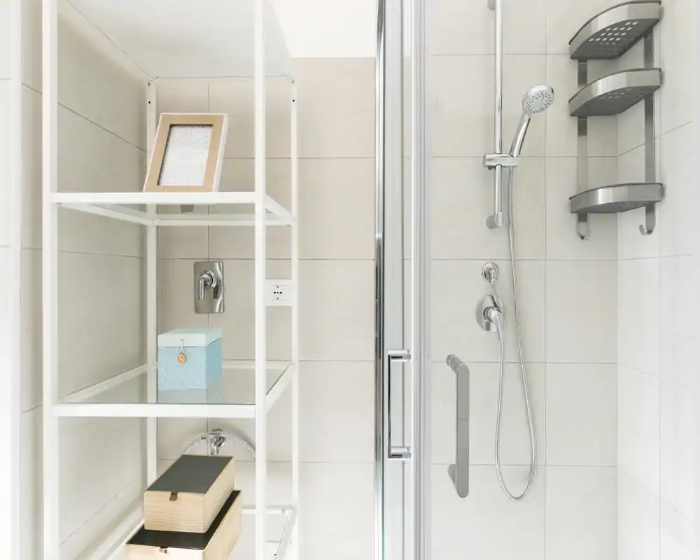 Rincón de baño moderno y luminoso con mampara de ducha de cristal, ducha de mano cromada y estanterías de pared.