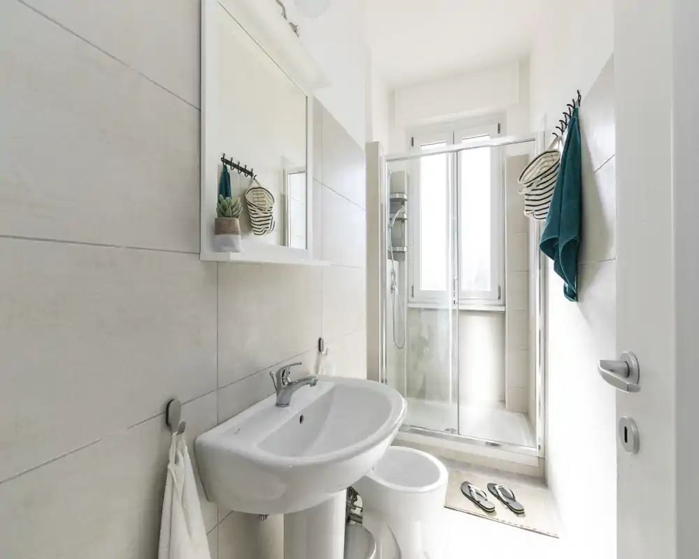 Baño moderno y luminoso con lavabo, inodoro y ducha con mampara de cristal frente a una ventana alta. Baldosas neutras y toallas verde azulado que aportan frescura.