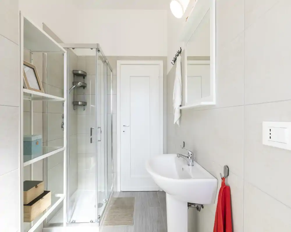 Baño moderno y luminoso con ducha de mampara de vidrio, lavabo pedestal y espejo, azulejos neutros y una toalla roja.