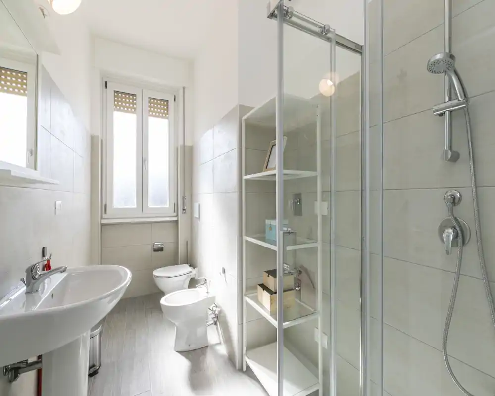 Baño moderno y luminoso con ducha de vidrio, lavabo, inodoro y bidé; azulejos neutros y luz natural desde una gran ventana.