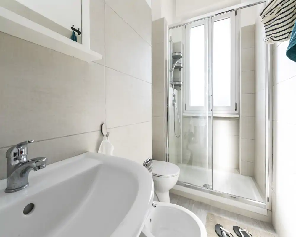 Baño moderno y luminoso con lavabo en primer plano, inodoro y ducha con mampara de cristal frente a una ventana grande.