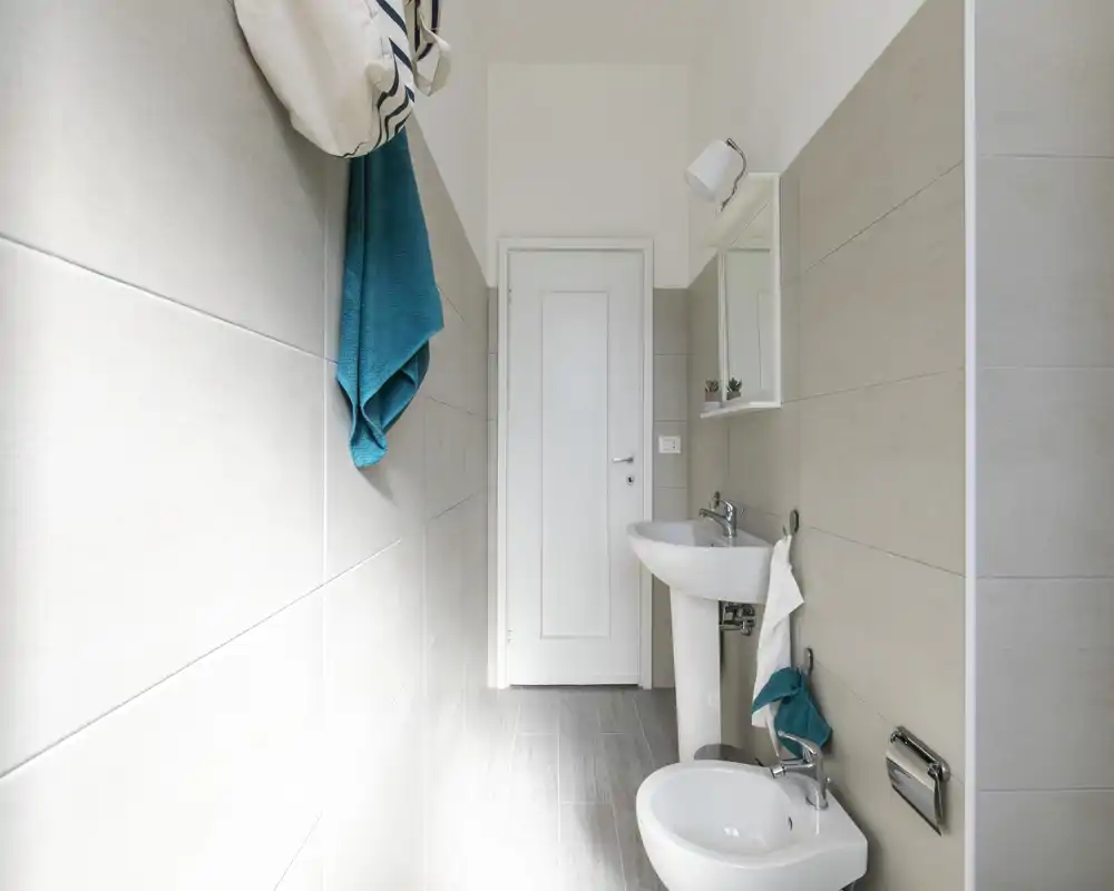 Baño moderno y luminoso con lavabo de pedestal, bidé y espejo de pared en un espacio estrecho con azulejos.