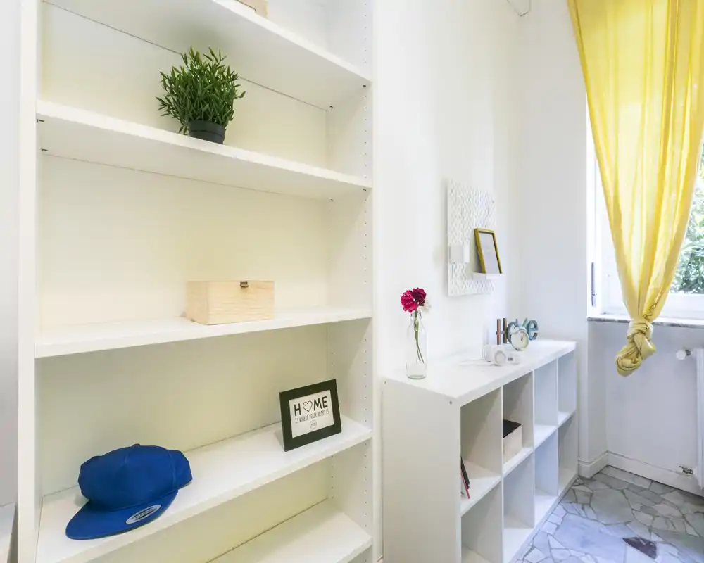Zona de estar luminosa y ordenada con estanterías blancas abiertas, objetos decorativos y una ventana soleada que crea una sensación aireada y minimalista.