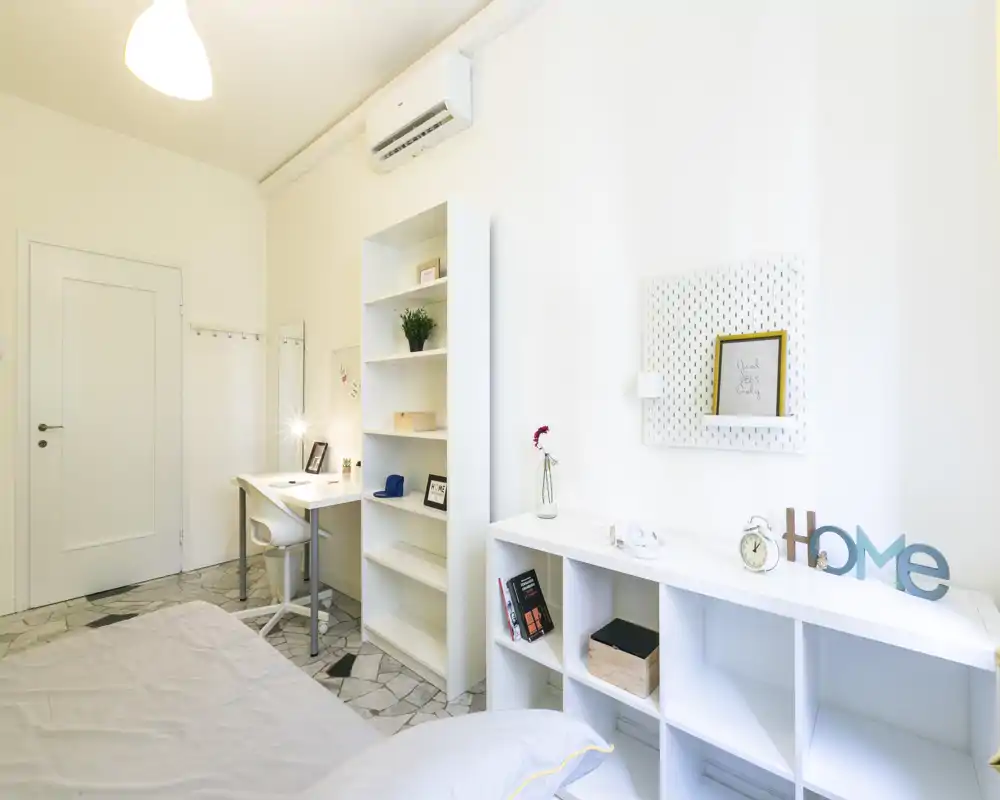 Dormitorio moderno y luminoso con cama visible en primer plano, estanterías ordenadas, zona de trabajo y decoración minimalista — ideal para alquiler.
