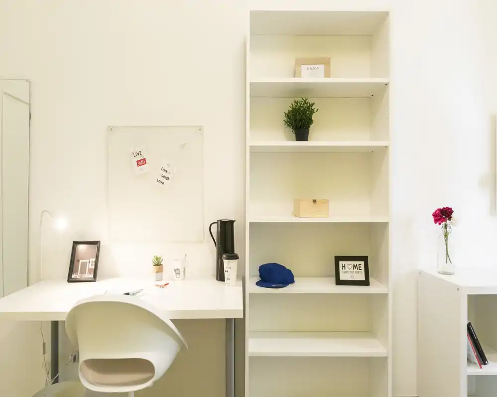 Rincón de trabajo luminoso y minimalista con mesa blanca, silla y estantería abierta — decoración ordenada, pequeña planta y marcos que crean un ambiente funcional y sereno.