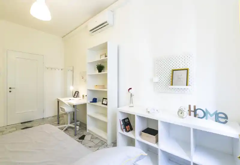 Camera da letto luminosa e moderna con letto visibile in primo piano, scaffalatura ordinata, scrivania per lavoro e decorazioni minimaliste — perfetta per affitti singoli.