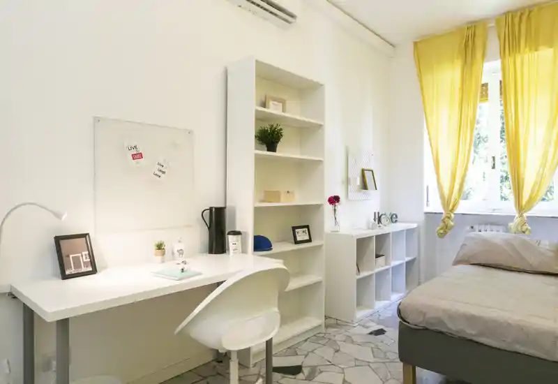 Camera da letto luminosa e ordinata con letto singolo vicino alla finestra con tende gialle, scrivania bianca e scaffalature che creano un angolo studio funzionale.