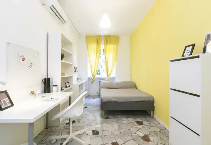 Camera da letto luminosa e ben arredata con letto matrimoniale contro una parete gialla, scrivania e scaffalatura sul lato opposto e luce naturale dalla finestra.