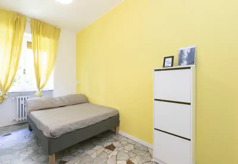 Camera da letto luminosa e semplice con letto matrimoniale contro una parete gialla, pavimento in piastrelle e luce naturale dalla finestra con tendine — ambiente pulito e accogliente.