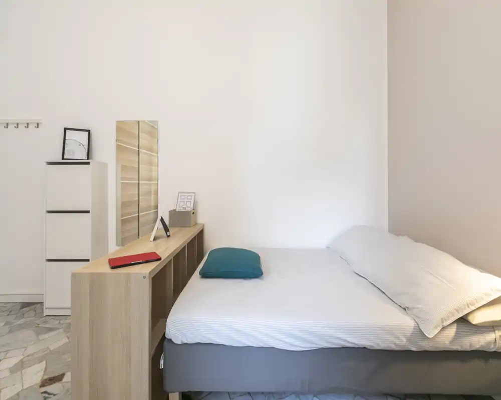 Dormitorio luminoso y minimalista con cama doble bien hecha, una estantería baja al pie de la cama y pequeños detalles decorativos para un aspecto moderno y sereno.