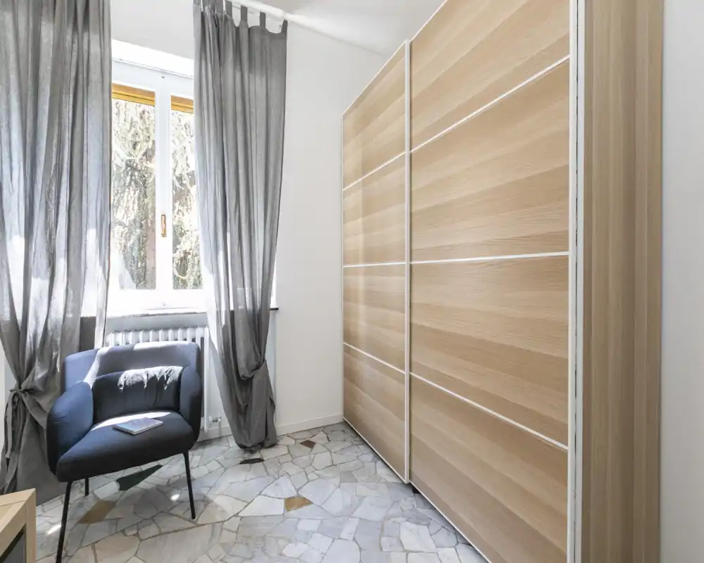 Rincón de dormitorio con un gran armario corredero de madera, un sillón cómodo junto a una ventana luminosa y cortinas grises largas; estilo moderno y sencillo, ideal para una persona o habitación de invitados.