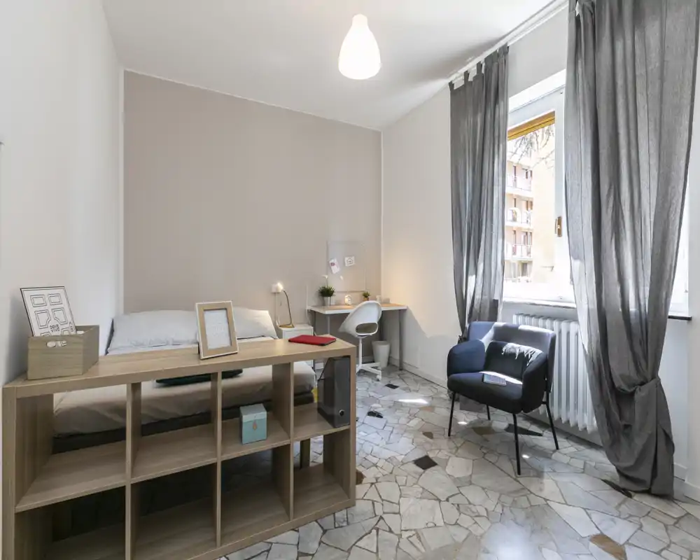 Dormitorio luminoso y bien preparado con cama individual, escritorio y un sillón acogedor junto a la ventana. La luz natural y los tonos neutros crean un ambiente tranquilo y agradable, ideal para anuncios.